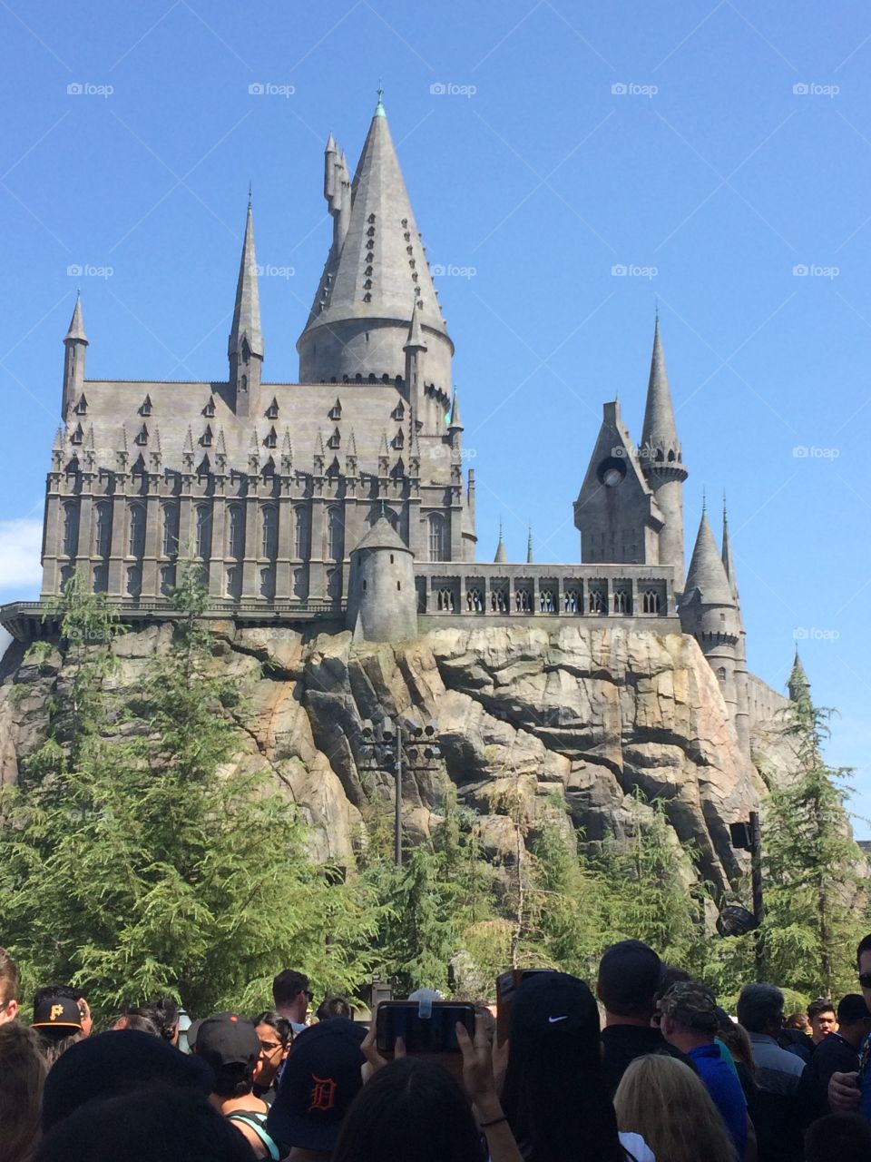 A beautiful sunlit shot of Universal Studios’ Hogwarts Castle atop a rocky platform. Y’all green trees feather the bottom of the hill.