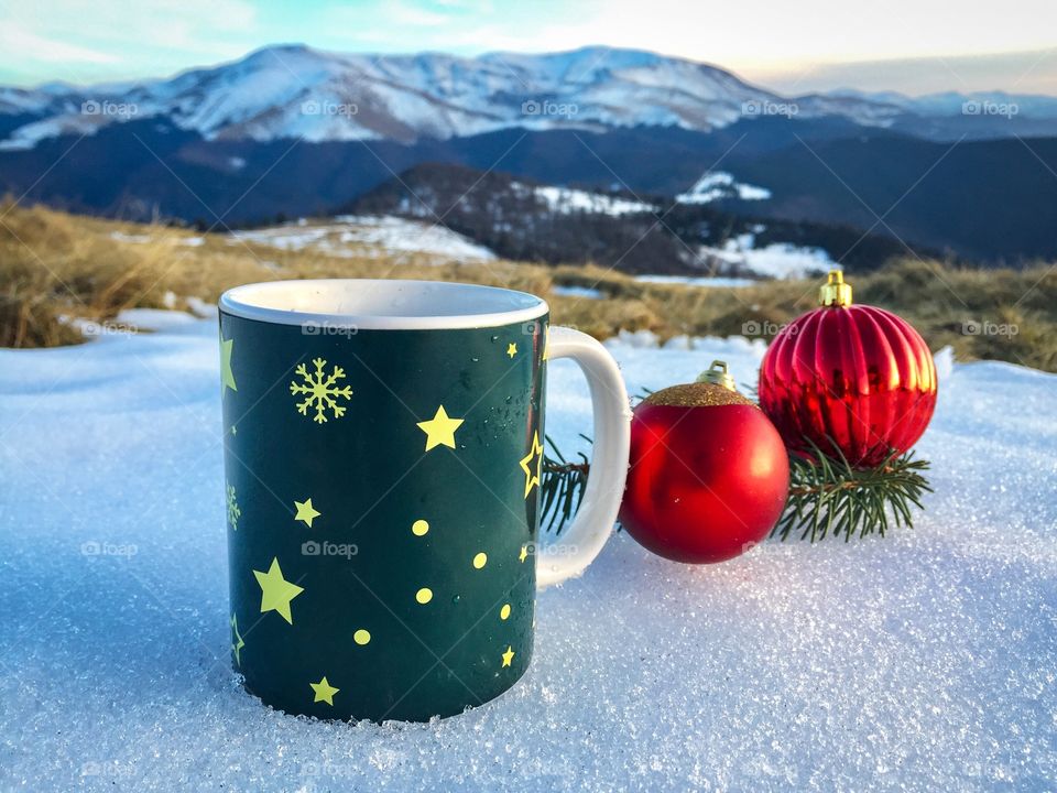 Snowflakes Christmas mug placed on snow with red globes and evegreen branches beside and snowy mountains in the background