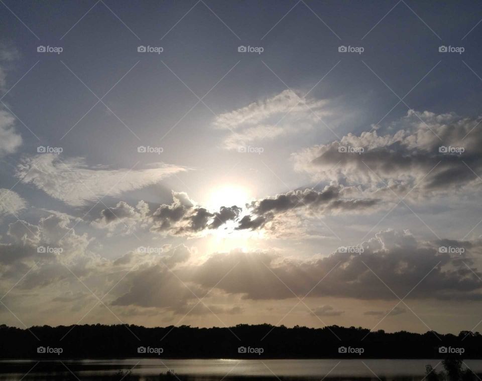 Descending Sun behind clouds with rays breaking through the bottom and shining in front of other clouds above distant trees and water.