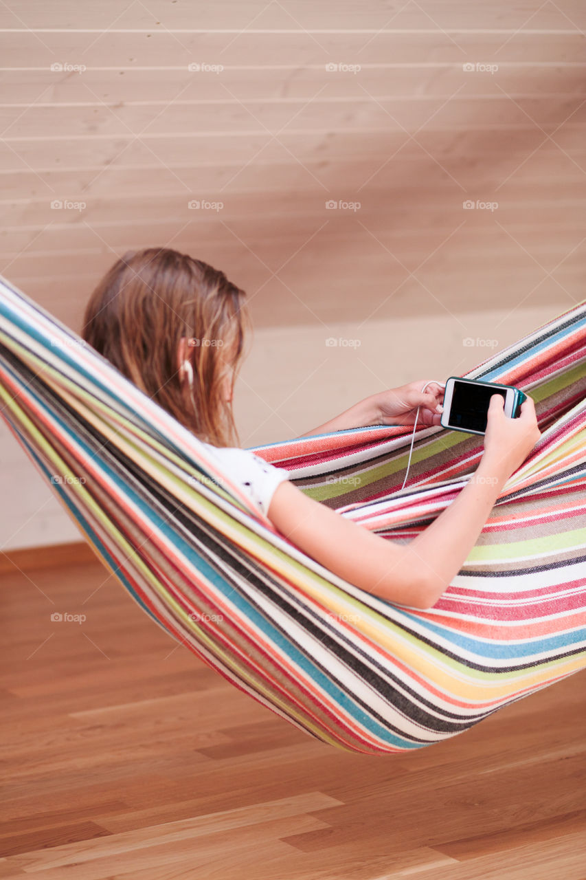 Teenage girl listening to music and chatting with friends on smartphone sitting in hammock at home