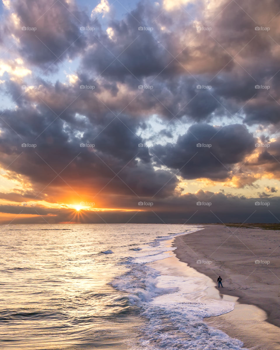 The sun poking through dark dramatic clouds over a beach, as a silhouette of a person stands in the sand watching the sunset.