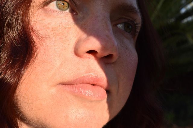 Women with natural and minimal makeup standing outdoors outside with wet hair. Women with red hair and green eyes standing next to tree taking a selfie. Sunlight shine on women hair and skin and making shadows. Women wearing a hat.
