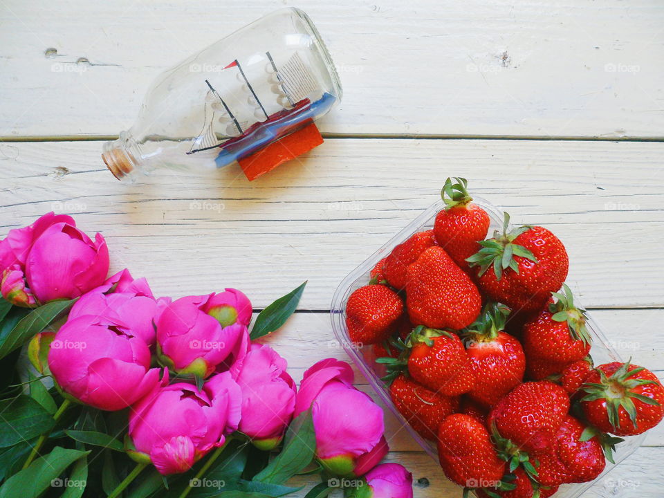 pink peony flowers, strawberry berries and souvenir in the form of a yacht in a bottle on a white background