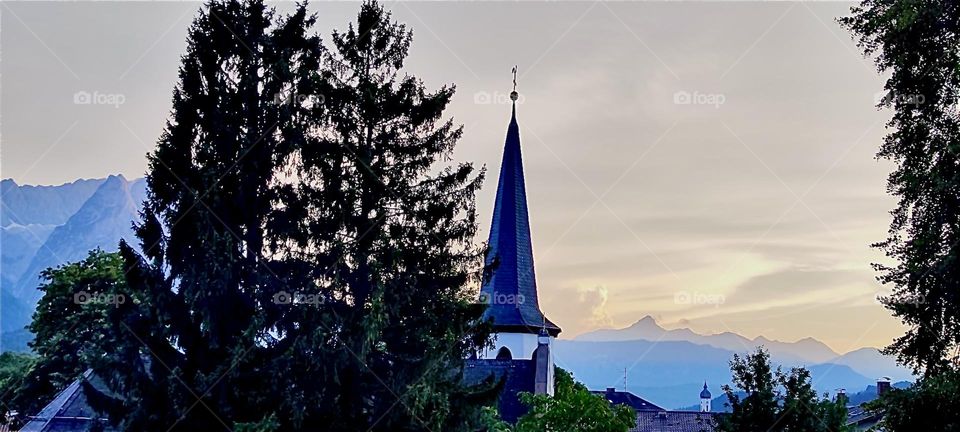 Silhouetted against a beautiful sunset is “Garmisch-Partenkirchen” with its forest and local church. The area is known for the “Zugspitze”, Germany’s tallest mountain in the “Alps” and is a popular ski resort in the winter. 2024. Hypnotic Productions