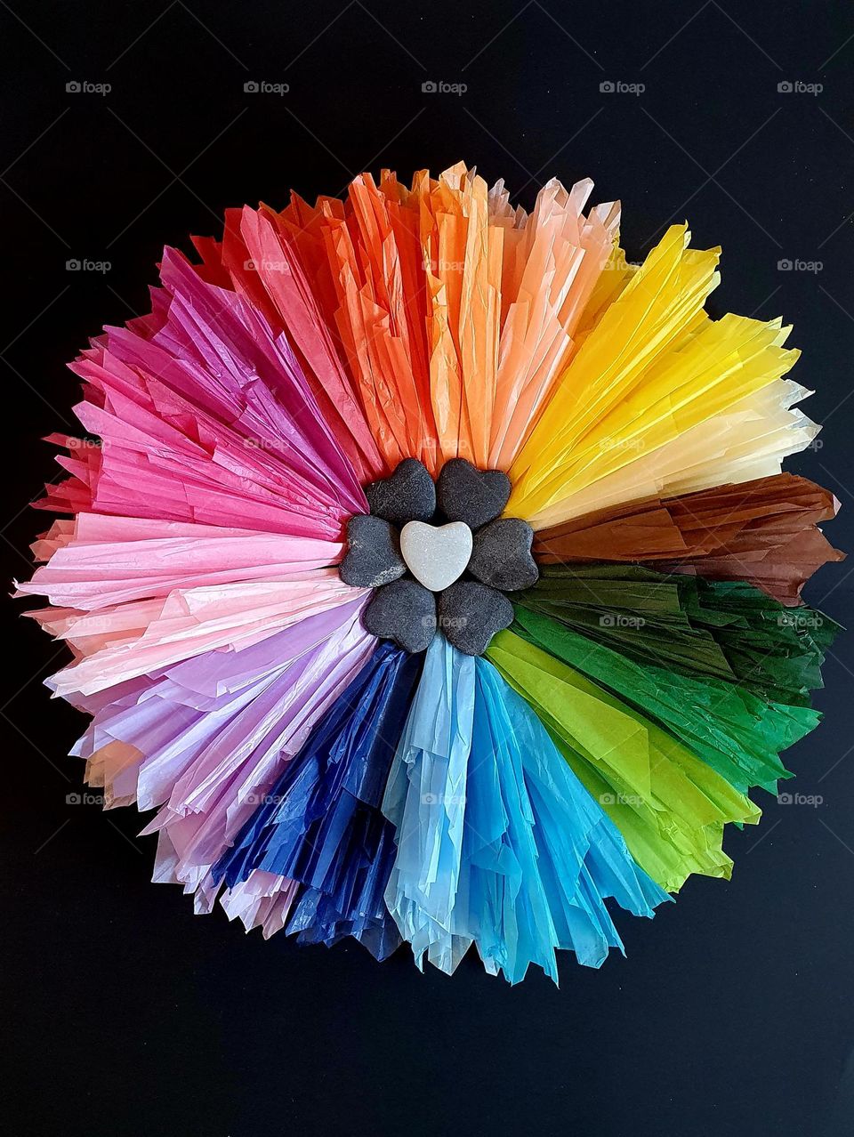 Flatlay composition of a multi-coloured flower made of silk papers and heart-shaped pebbles placed on black paper, creating a colour explosion