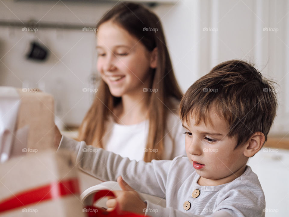 Cute toddler boy in grey pajamas smile in kitchen and packing Christmas gifts with his sister