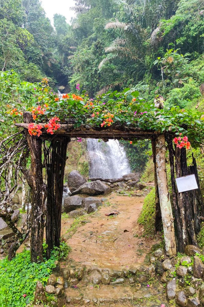 A natural gate surrounded by beautiful flowers with a waterfall as a backdrop in the middle of a green forest