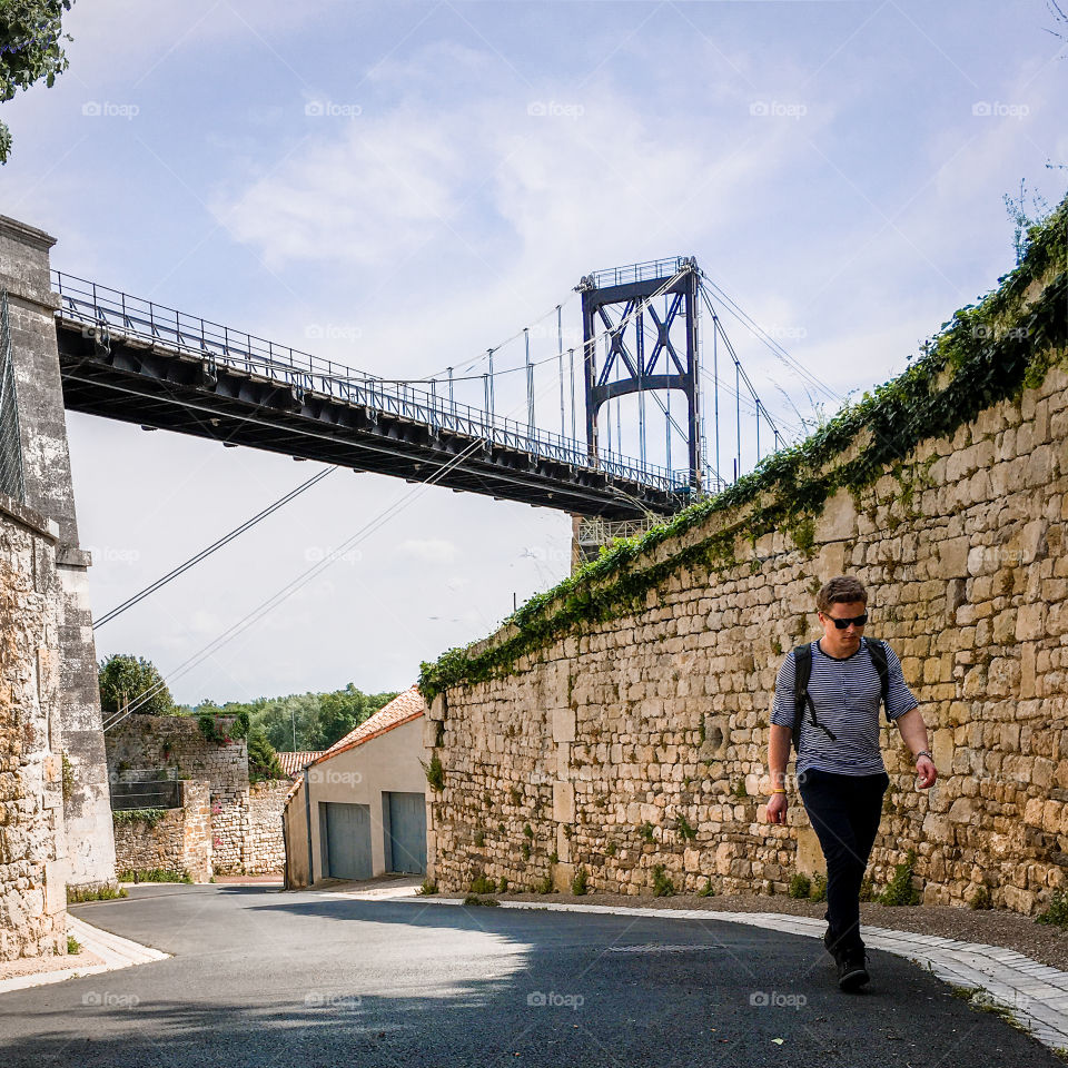 Young man walking on the pavement with a large bridge in the background.