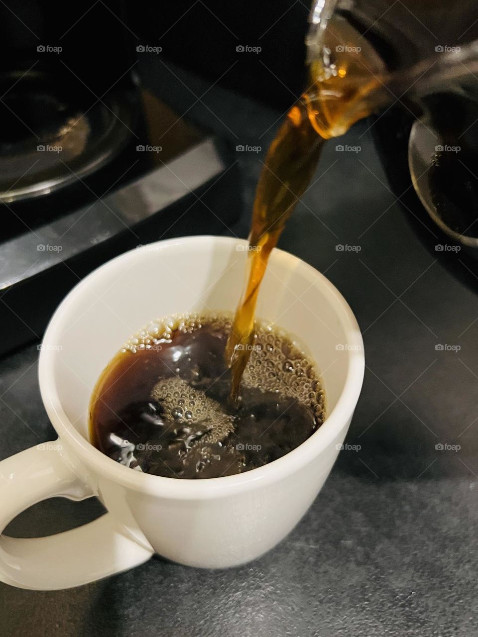 Overhead closeup of white coffee cup. Coffee is pouring from above into the half-filled mug in front of a black drip coffee maker. Setting is a home kitchen, a black laminate  counter with black appliances.