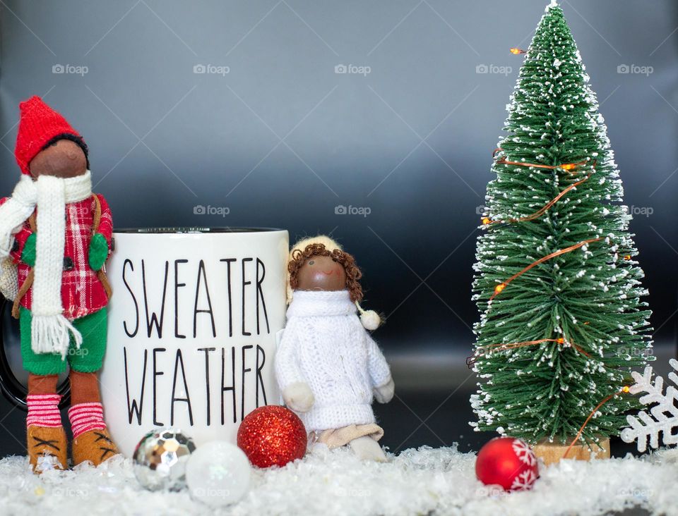 A cute holiday scene with a mug,dolls,ornaments snow and a lit tree