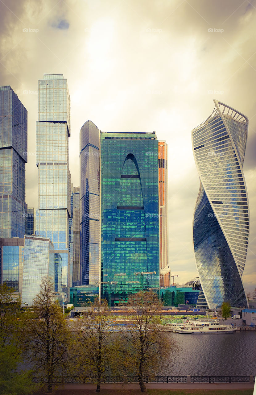 Russia, Moscow, Moscow-City district. Spring cityscape with modern high-rise buildings. View of skyscrapers and the Moskva River with pleasure boats and a promenade with trees with blossoming leaves
